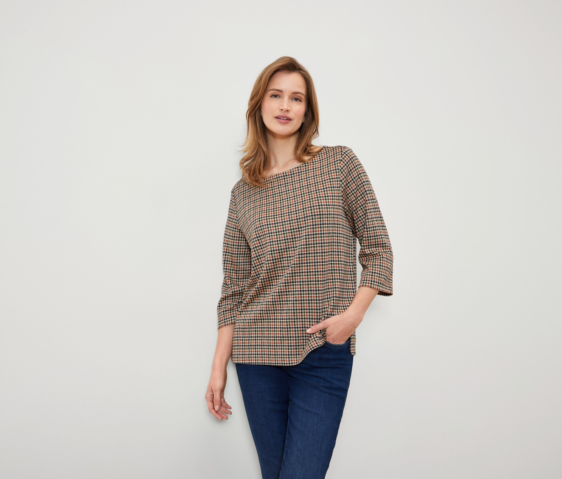 Žena s hnedými vlasmi pózuje v kockovanej hnedej blúzke a tmavomodrých džínsoch.