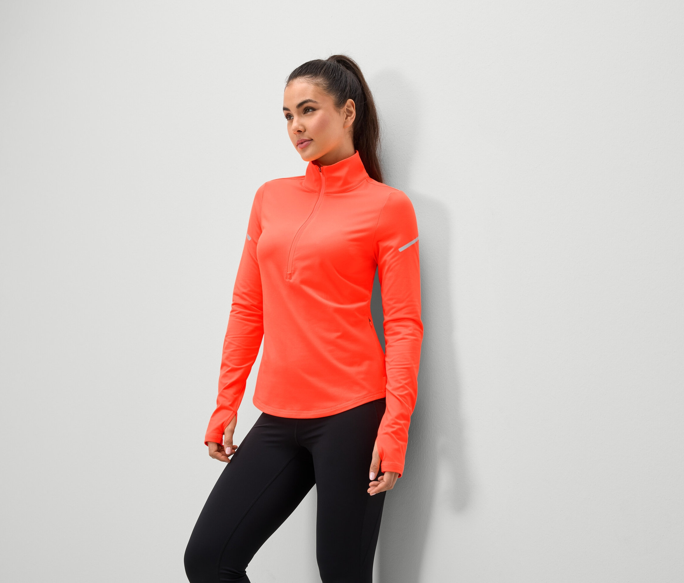 Žena v koralovej športovej bunde s čiernymi legínami stojí pri stene.