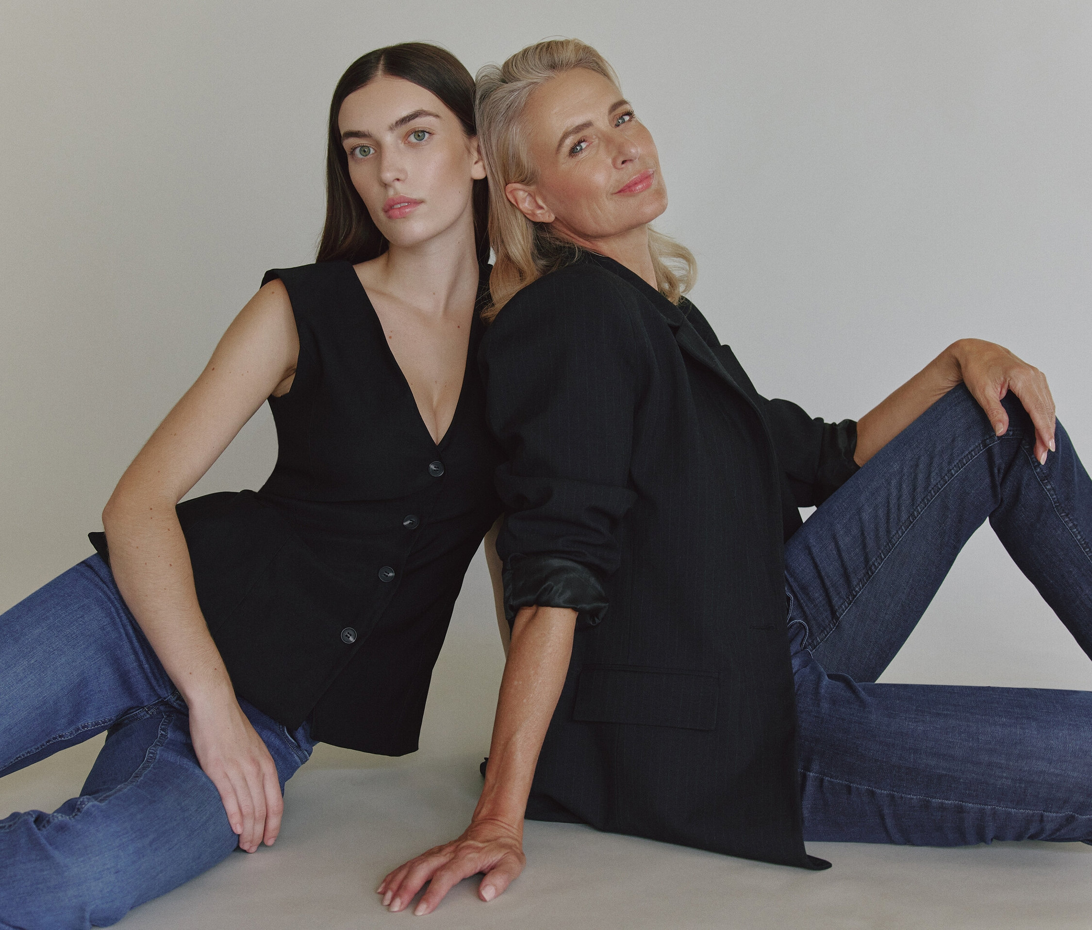 Dve ženy sedia chrbtom k sebe na podlahe. Jedna má na sebe čierne sako a džínsy, druhá čiernu vestu a džínsy.