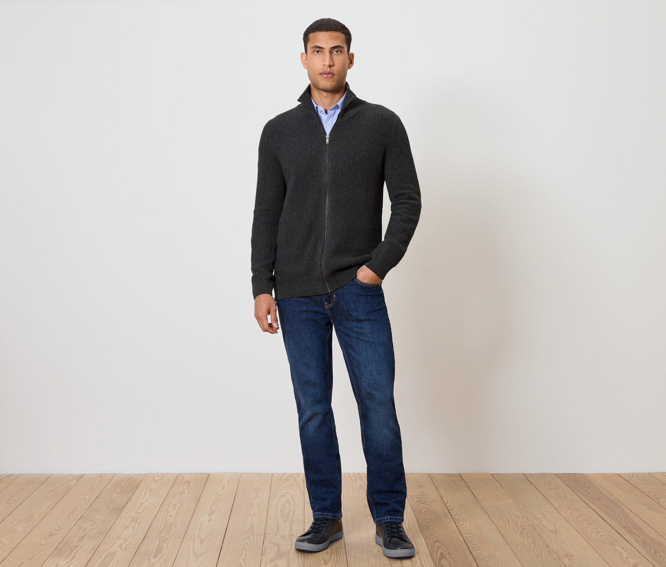 Muž stojí a má na sebe pletený sveter, antracitový s melírom, rifle a topánky.