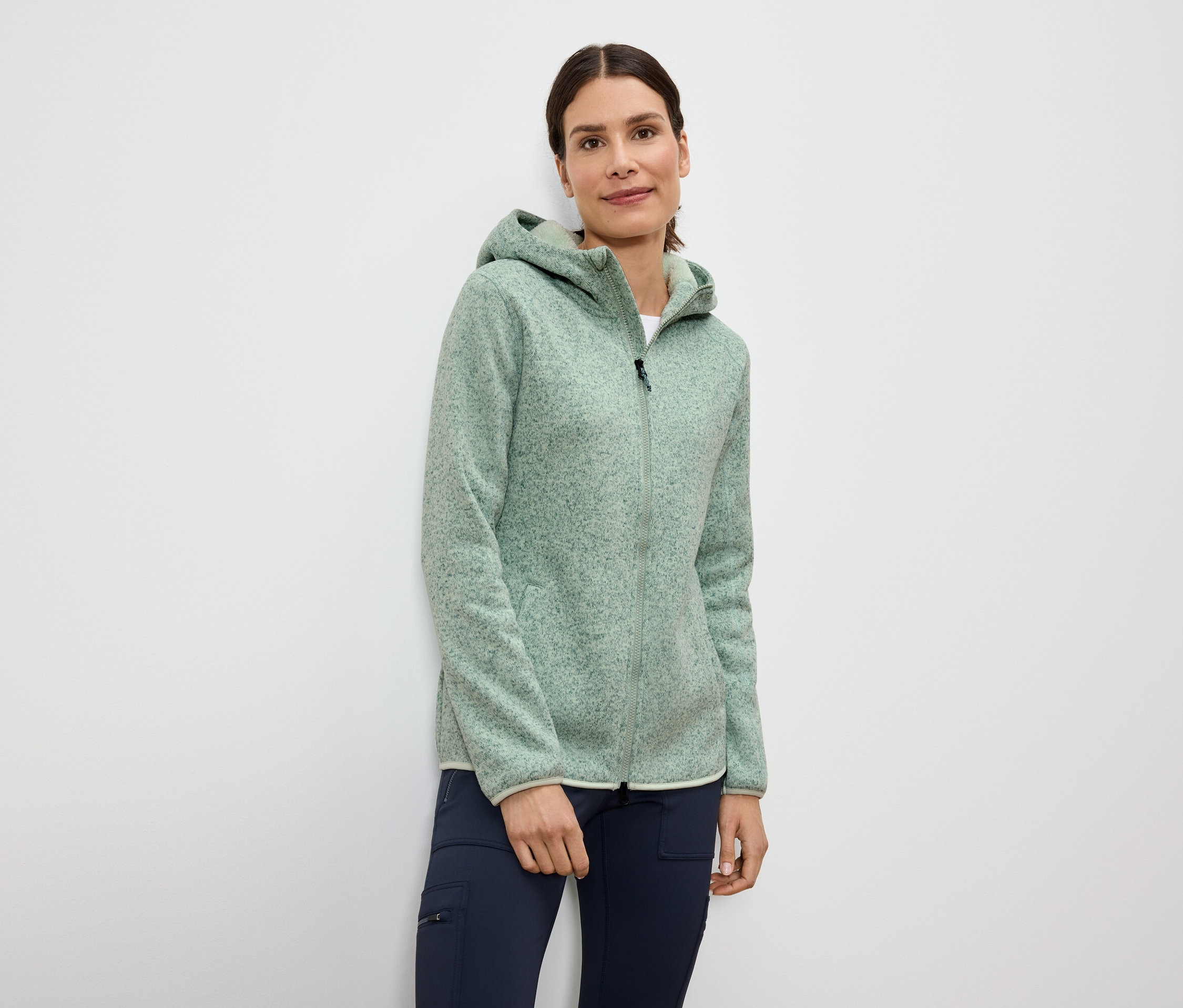 Žena má na sebe svetlozelenú flisovú bundu na zips s kapucňou a tmavomodré nohavice.