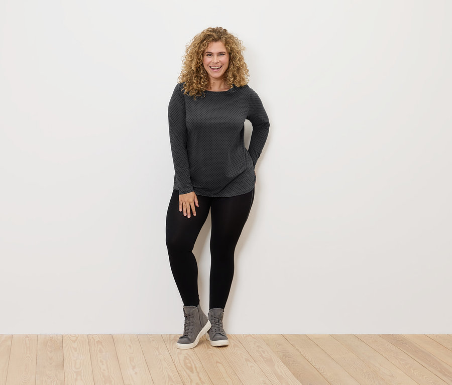 Žena s kučeravými vlasmi stojí a usmieva sa, má na sebe čierne legíny, sivé topánky a sivo-čierne tričko.