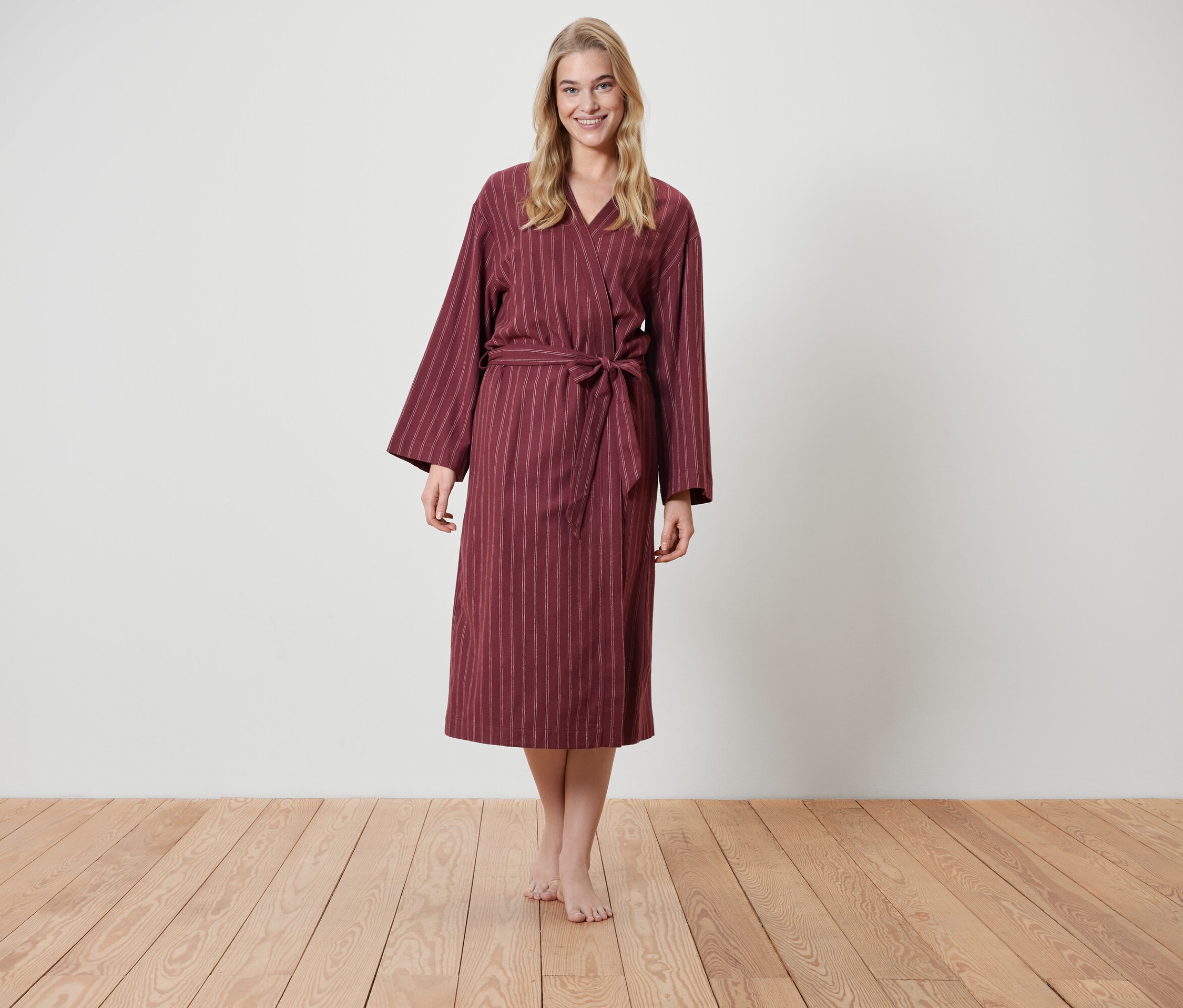 Blondínka stojí na drevenej podlahe a má na sebe vínový župan s prúžkami.