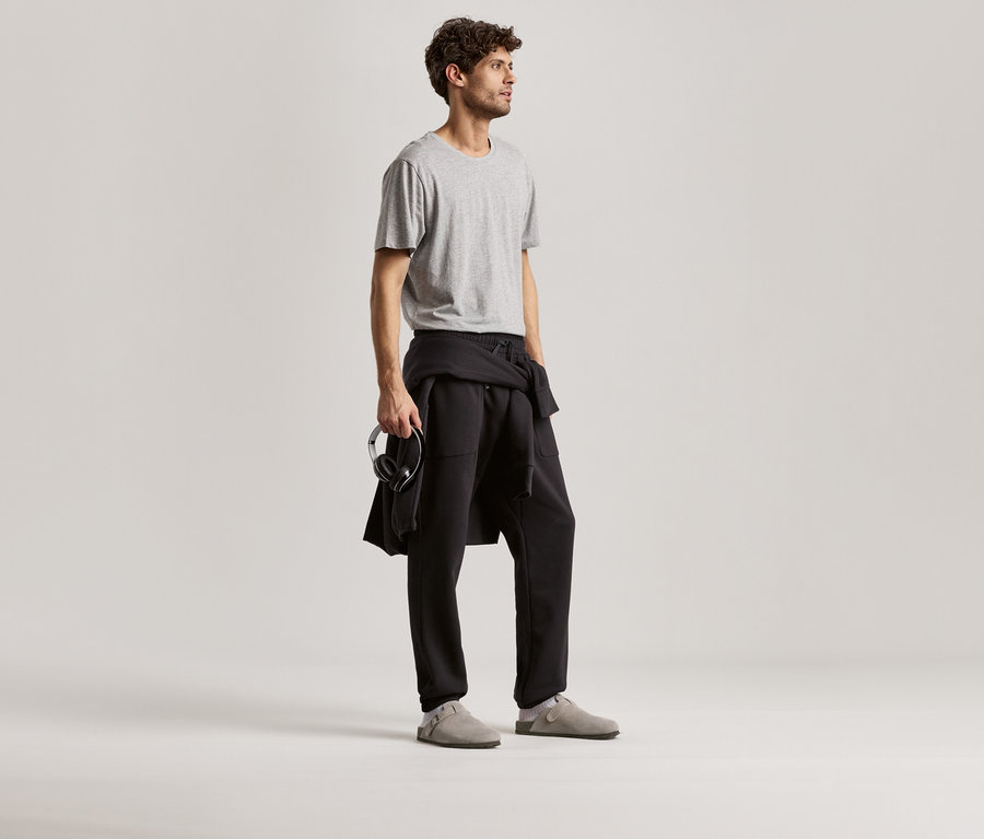 Muž má na sebe sivé tričko a čierne teplákové nohavice. V ruke drží slúchadlá.