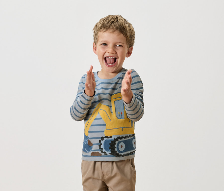 Chlapec s blond vlasmi tlieska rukami, oblečený v modro-pruhovanom tričku s dlhým rukávom so žltým buldozérom.