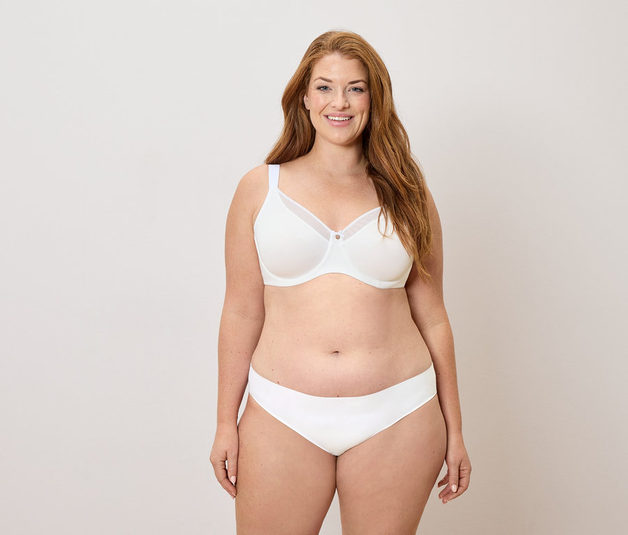 Žena s dlhými ryšavými vlasmi v bielej podprsenke a nohavičkách sa usmieva.