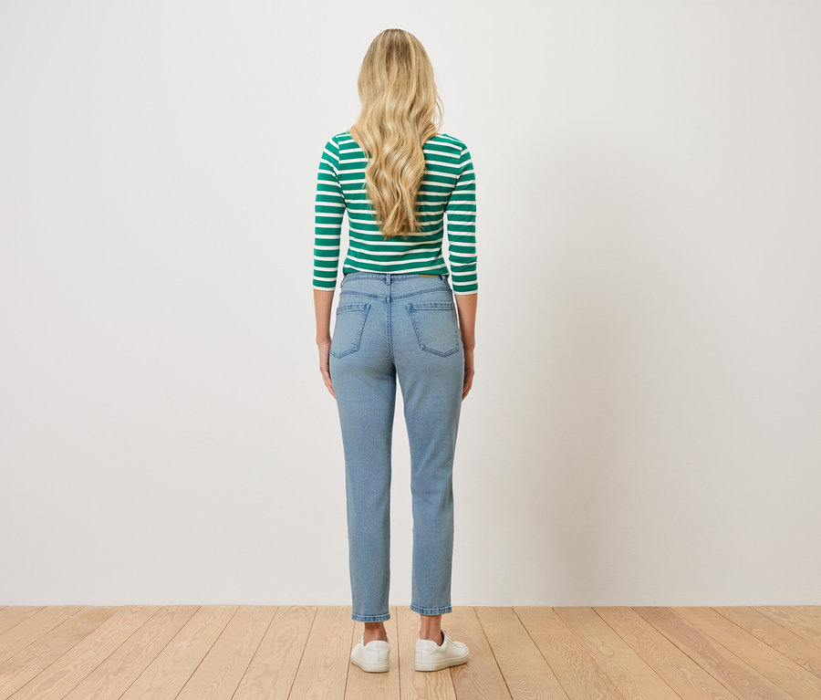 Pohľad zozadu na ženu s blond vlnitými vlasmi, oblečenú v zeleno-bielom pruhovanom tope, svetlomodrých džínsoch a bielych teniskách.