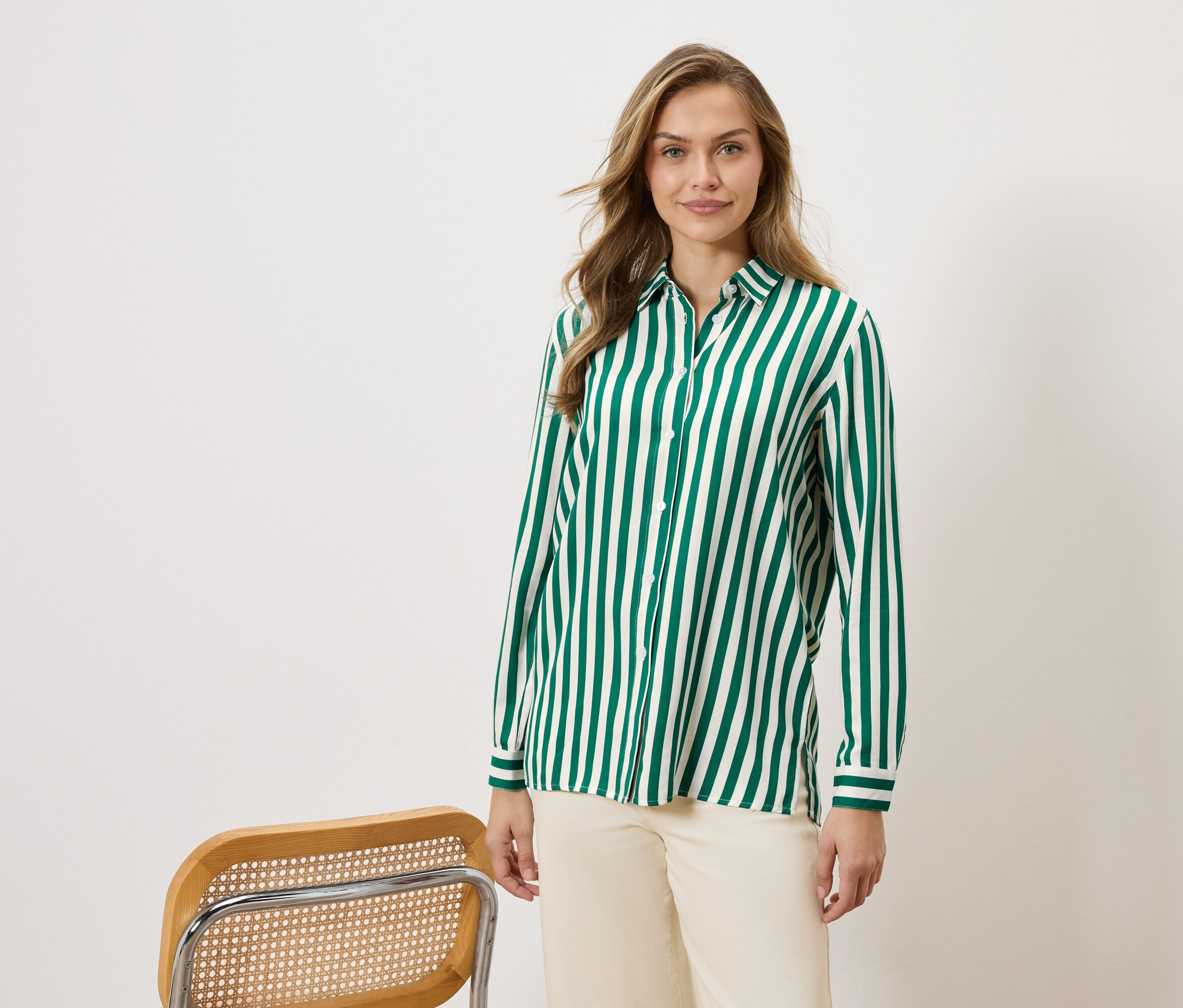 Žena má na sebe zeleno-bielu pruhovanú košeľu a béžové nohavice, vedľa stoličky.