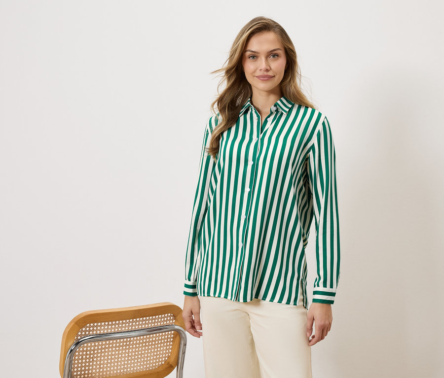 Žena má na sebe zeleno-bielu pruhovanú košeľu a béžové nohavice, vedľa stoličky.