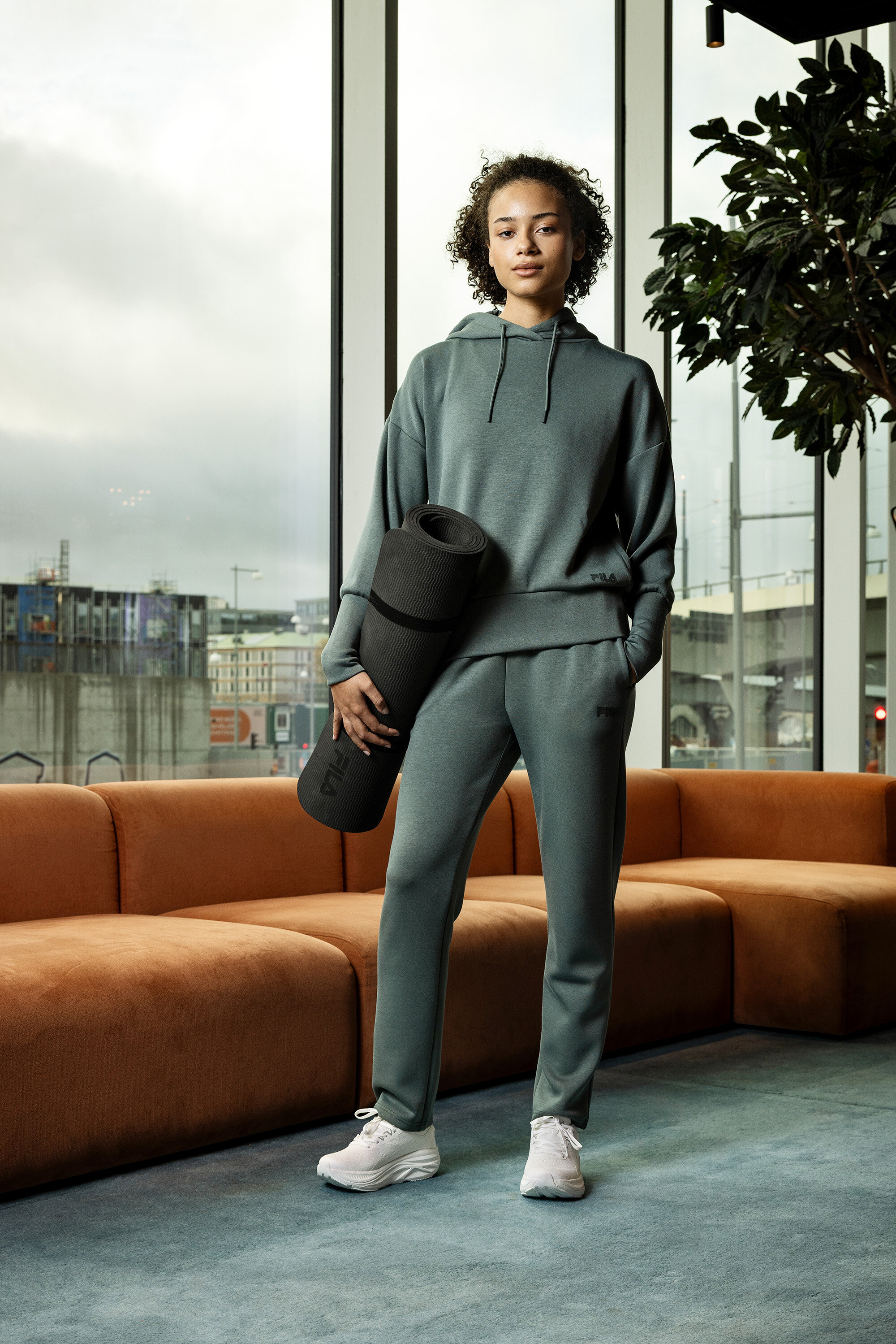 Mladá žena v sivom športovom oblečení drží zrolovanú podložku na cvičenie jogy pred oknom.