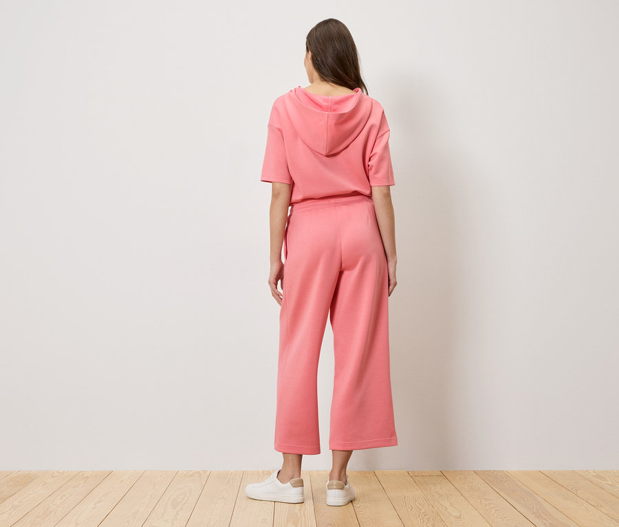 Žena stojí chrbtom k fotoaparátu a má na sebe ružovú mikinu s kapucňou a culottes nohavice.