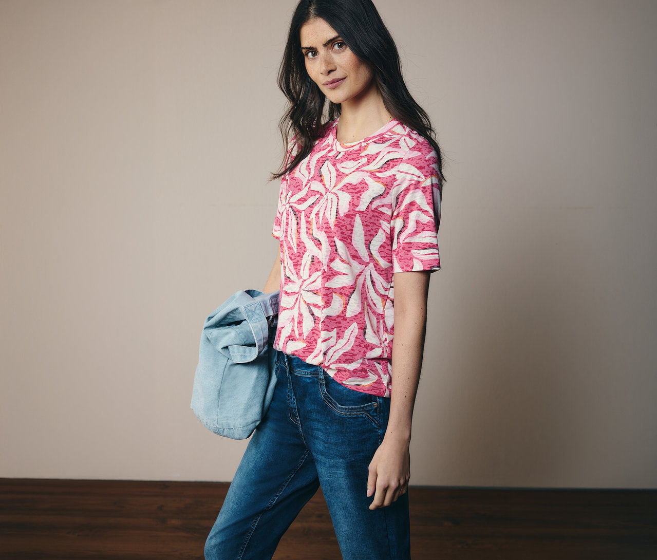 Žena má na sebe ružové tričko s krátkym rukávom CECIL so vzorom a džínsy. Na ramene má džínsovú bundu.