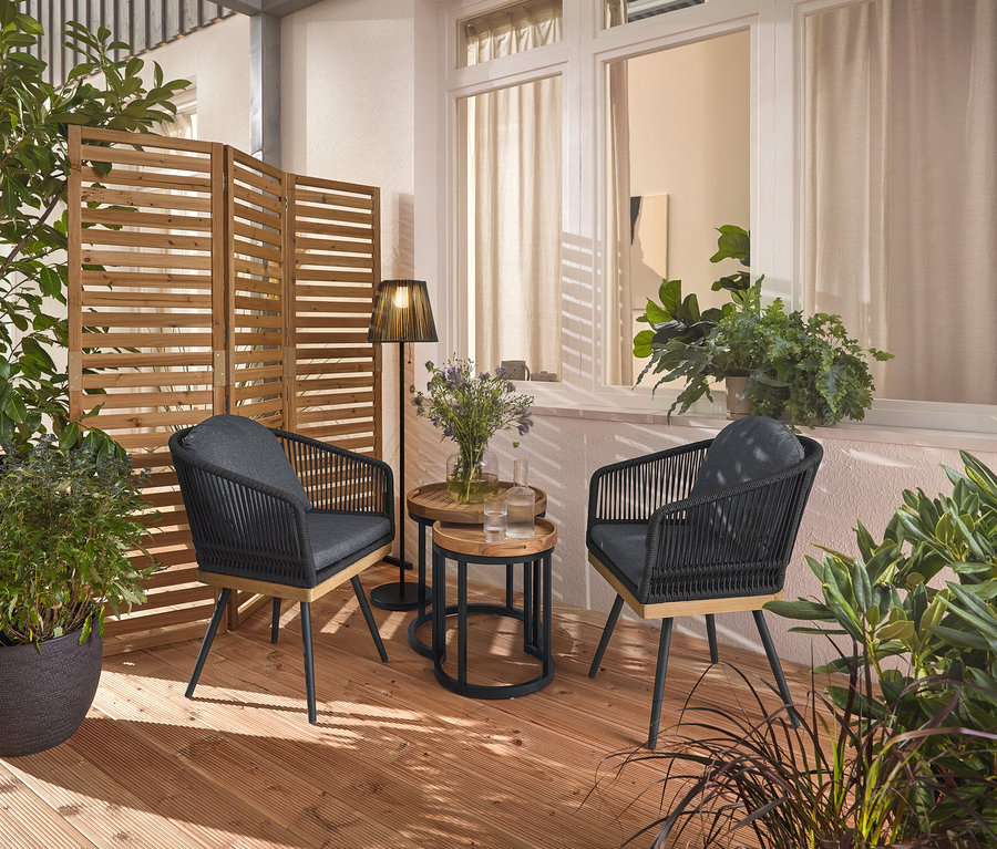 Dve otočné záhradné stoličky s textilným výpletom stoja na drevenej terase, medzi nimi dva záhradné stolíky s odnímateľnými podnosmi, Ø cca 40 cm a 50 cm. V pozadí flexibilný paraván a akumulátorová solárna stojacia lampa.