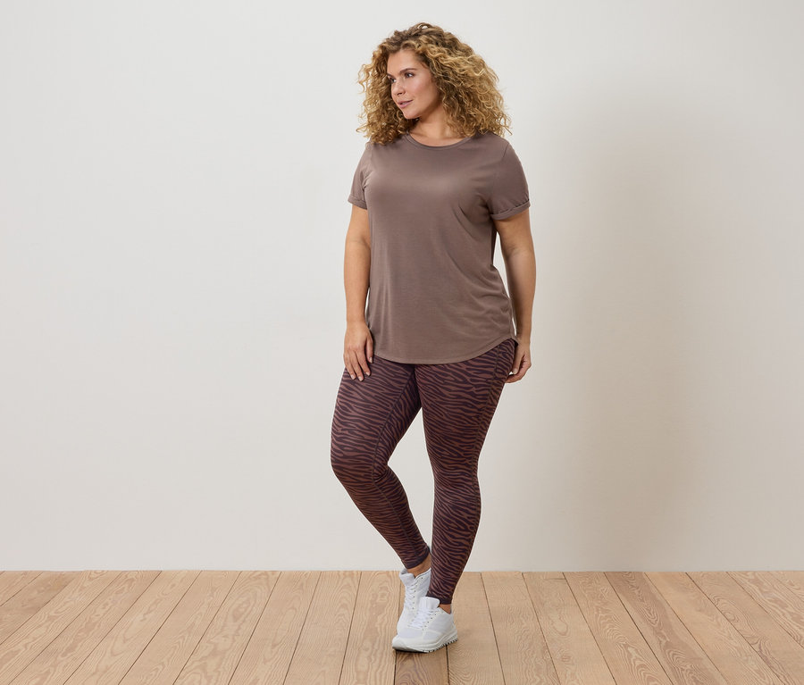 Žena s kučeravými vlasmi, hnedým tričkom a vzorovanými legínami pózuje pred stenou.