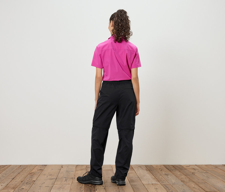 Pohľad zozadu na ženu s kučeravými vlasmi v ružovej košeli a čiernych nohaviciach so zipsom a čiernych topánkach.