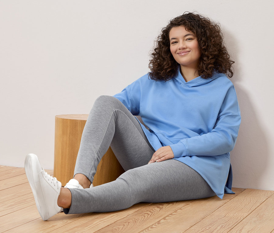 Žena s kučeravými vlasmi sedí na zemi, má na sebe svetlomodrú mikinu s kapucňou, sivé legíny a biele tenisky.