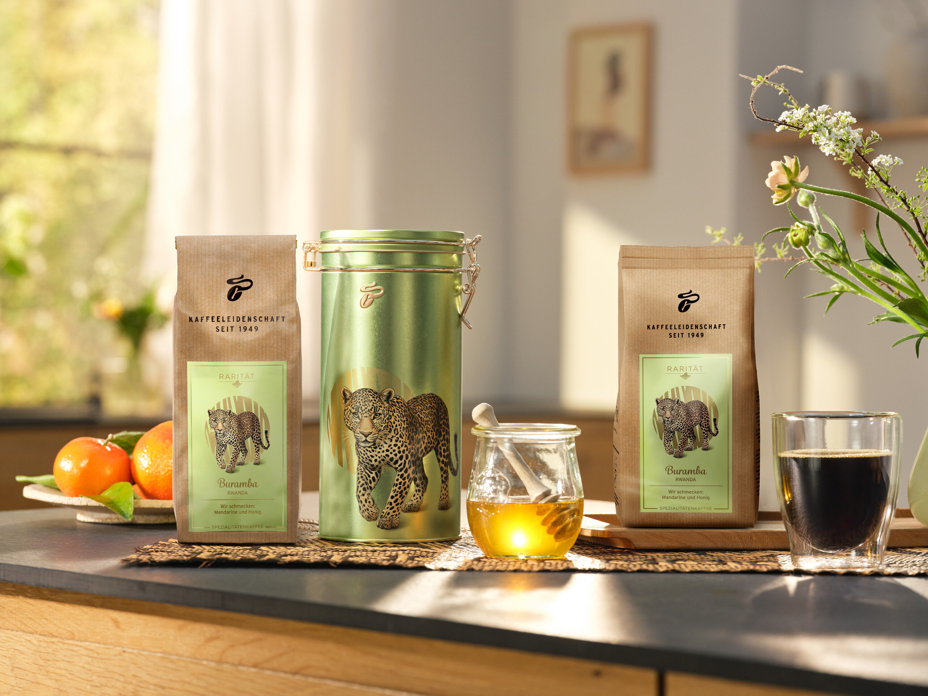Na kuchynskej linke stojí dóza na kávu a dve balenia »Buramba Rwanda« vrátane dózy na kávu vedľa šálky kávy a medu.