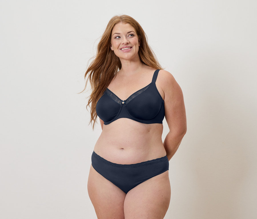 Žena s dlhými ryšavými vlasmi má na sebe námornícku modrú podprsenku a nohavičky a usmieva sa.