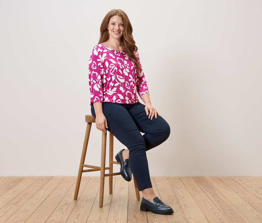 Žena sedí na stoličke, má na sebe ružovú blúzku s kvetinovým vzorom, modré nohavice a kožené topánky.