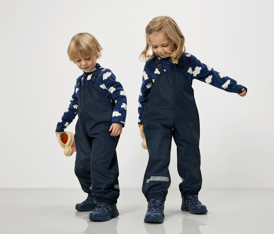 Dve deti v tmavomodrých kombinézach a modro-bielych obláčikových svetroch stoja na bielom pozadí.