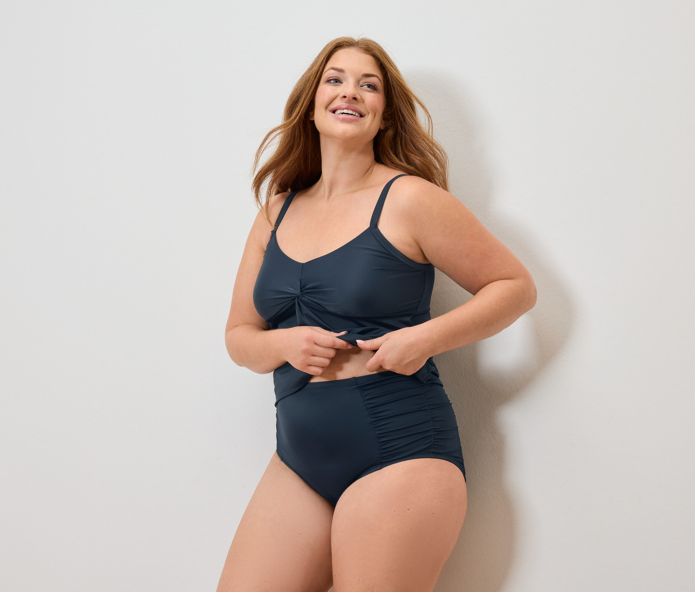 Žena s dlhými červenými vlasmi v tmavomodrých plavkách sa usmieva.