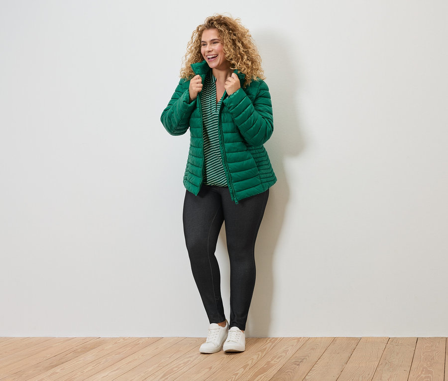 Žena s kučeravými vlasmi v zelenej prešívanej bunde, pásikavej blúzke, tmavých legínach a bielych teniskách sa usmieva.
