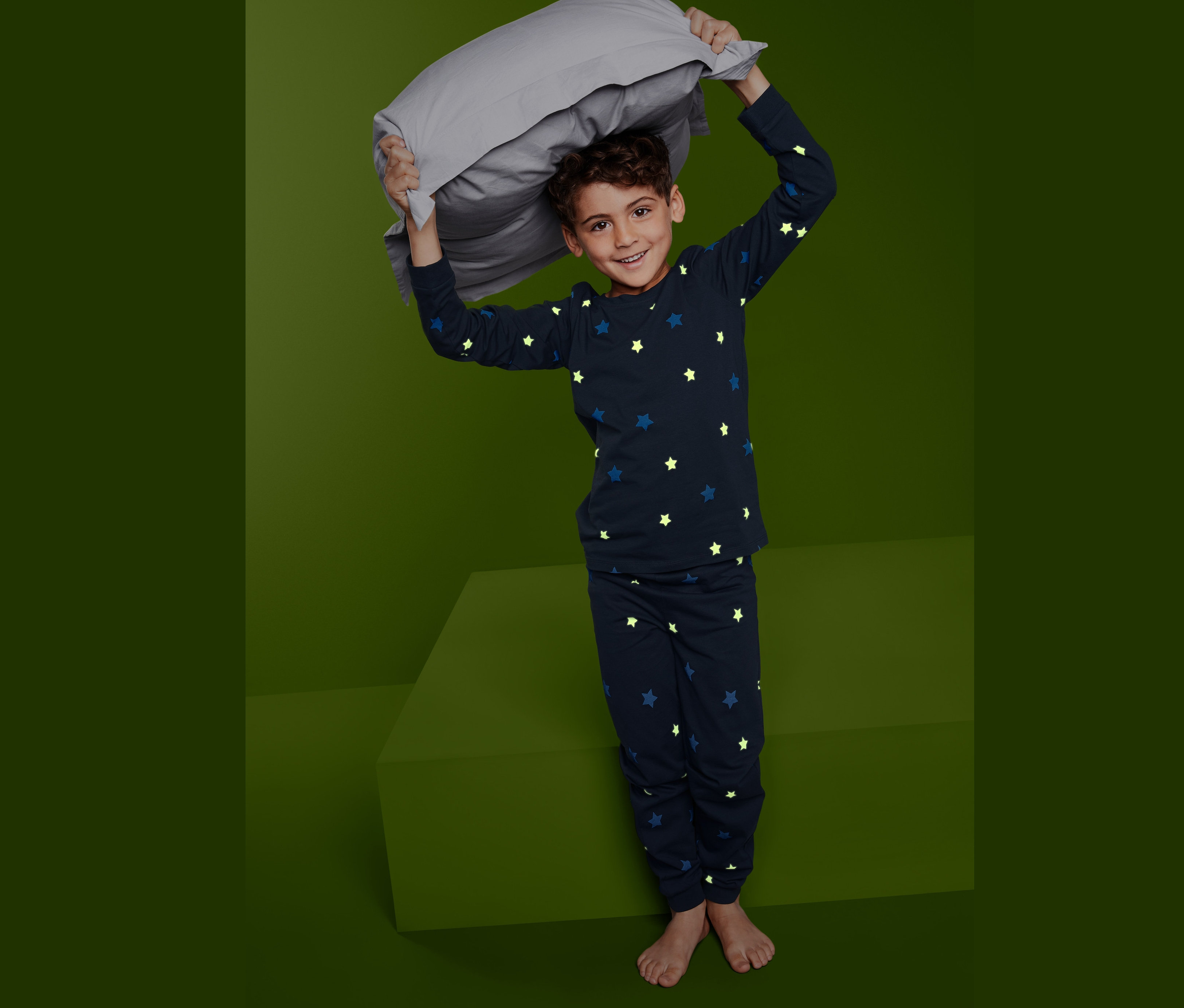 Chlapec drží nad hlavou sivý vankúš, oblečený do tmavomodrého pyžama »Glow in the dark« s hviezdami.