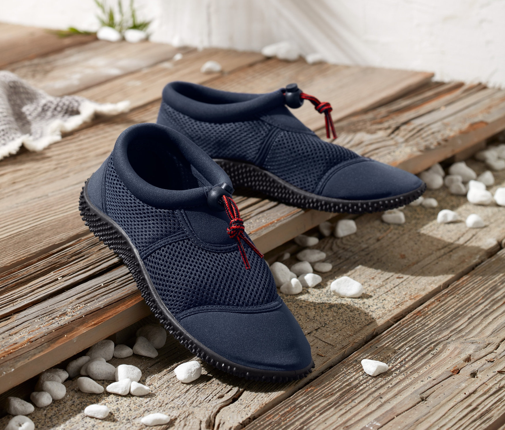 Dva páry modrých topánok na kúpanie ležia na drevenom povrchu posypanom kamienkami.