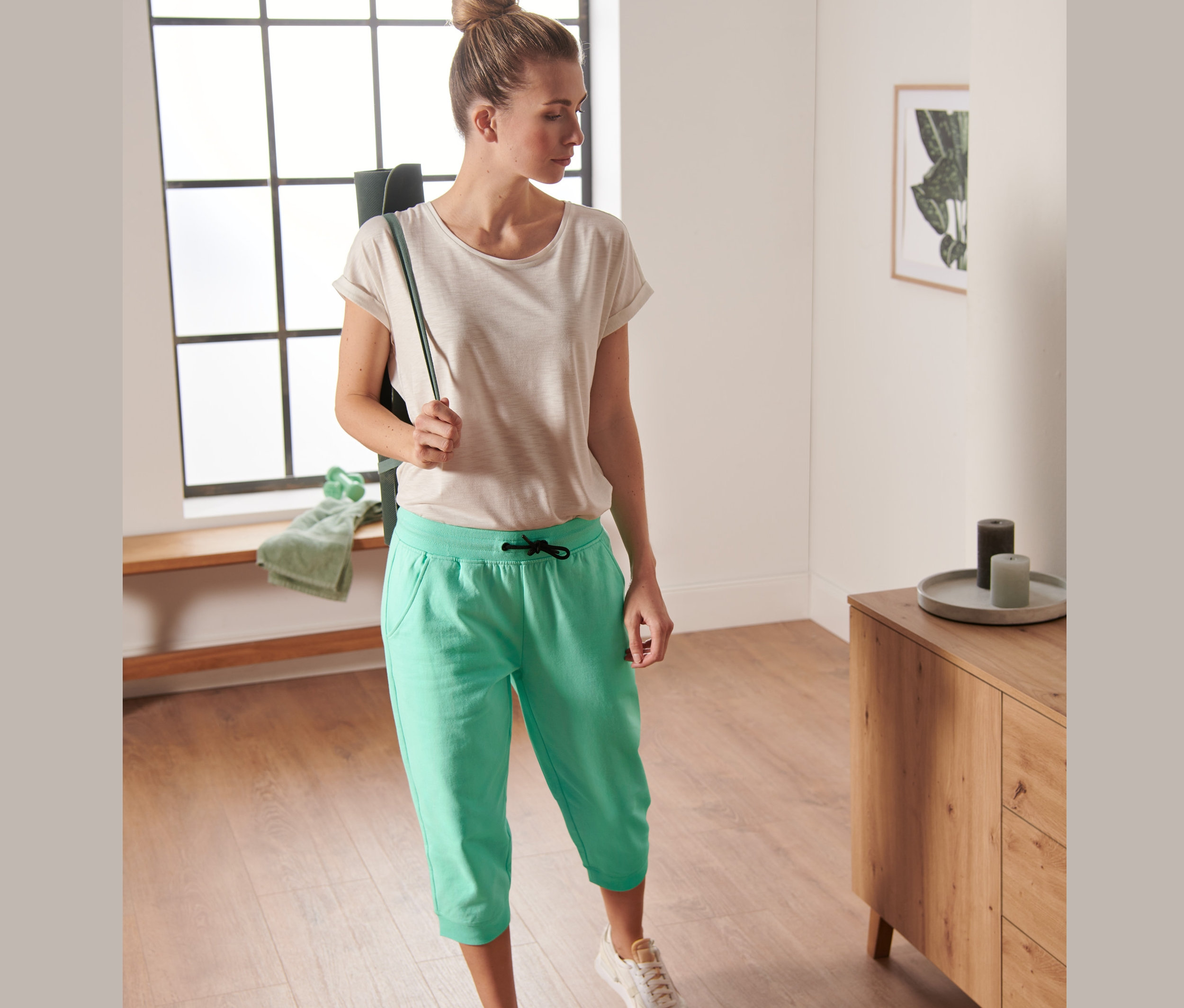 Žena má cez rameno podložku na cvičenie a na sebe má trojštvrťové športové nohavice, mätové.