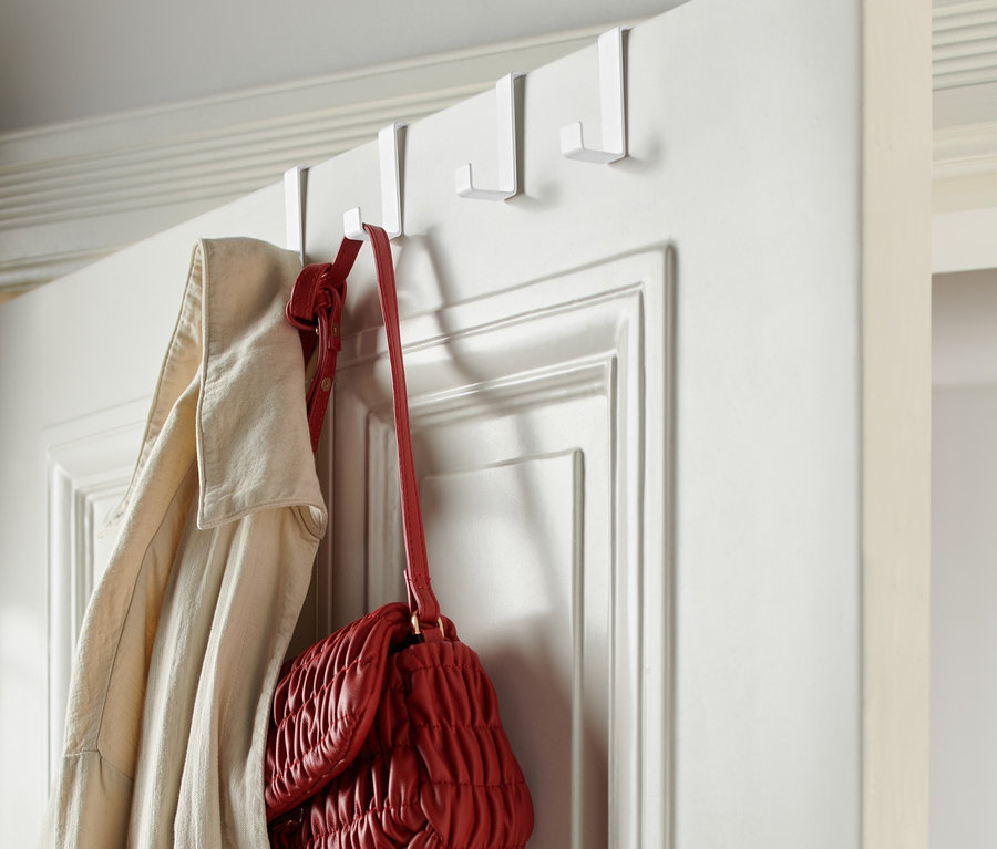Na bielych dverách visia 4 biele háčiky na dvere, na ktorých visí béžový kabát a červená kabelka.