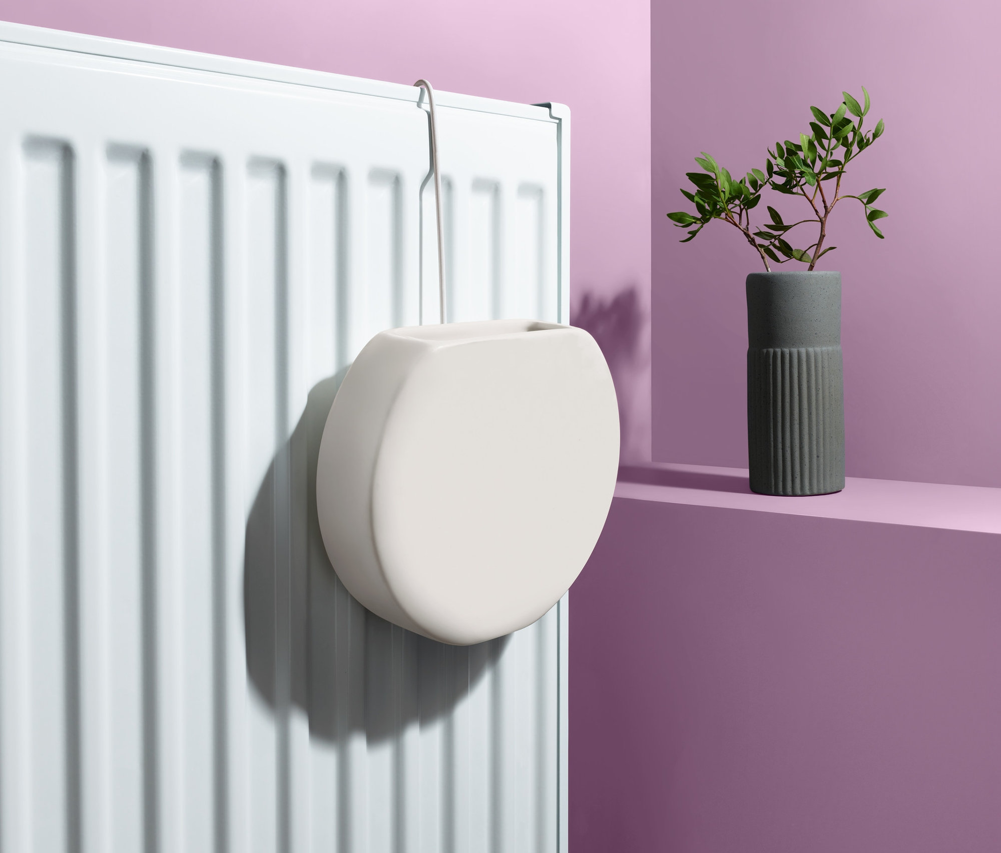 Biely zvlhčovač vzduchu na radiátor zavesený na bielom radiátore. Vedľa stojí váza so zelenými listami.