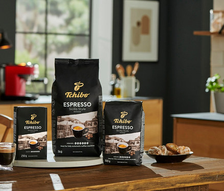 Tri balenia kávy Tchibo Espresso Sicilia Style (1 kg, 500 g, 250 g) stoja na drevenom stole vedľa šálky espressa a taniera sušienok.