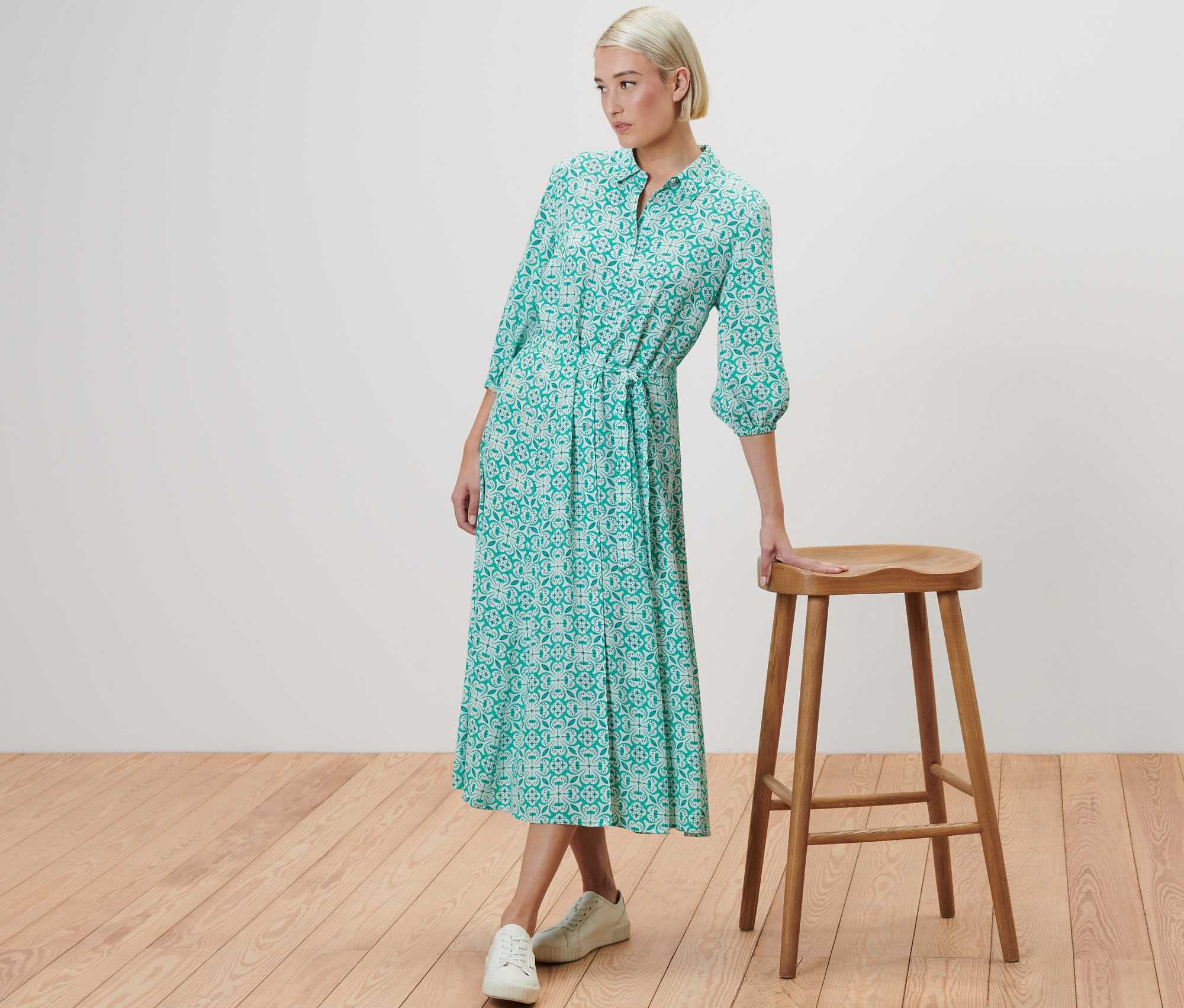 Žena pózuje vedľa drevenej stoličky a má na sebe košeľové šaty s celoplošnou potlačou.