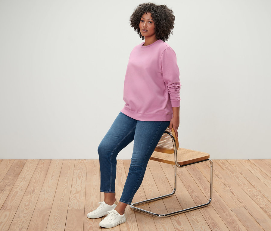 Žena sedí opretá o stoličku, má na sebe ružovú mikinu, tregínsy »Fit Nelly« a biele tenisky.