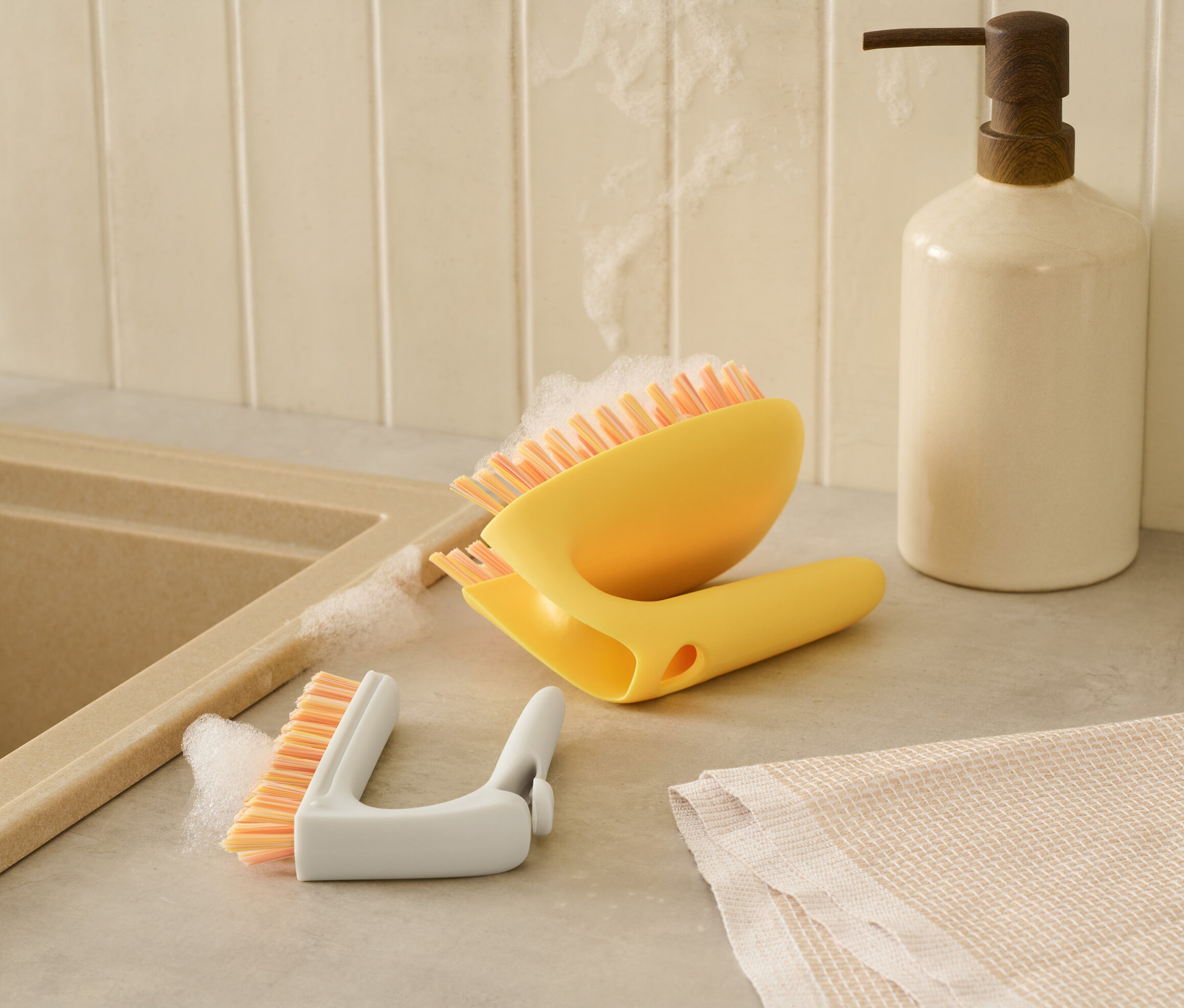 Žltá kefa na dlaždice 2 v 1 a biela kefa na detaily ležia vedľa seba na pulte pri umývadle.