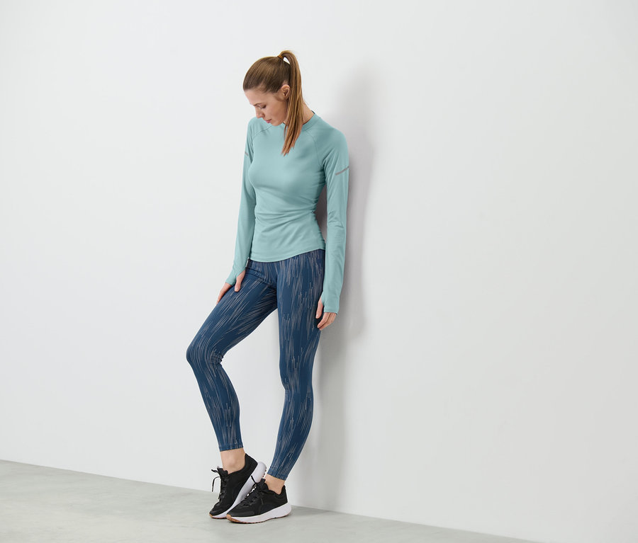Žena opretá o stenu má na sebe športové legínsy, reflexné v striebornej farbe, a top s dlhým rukávom.