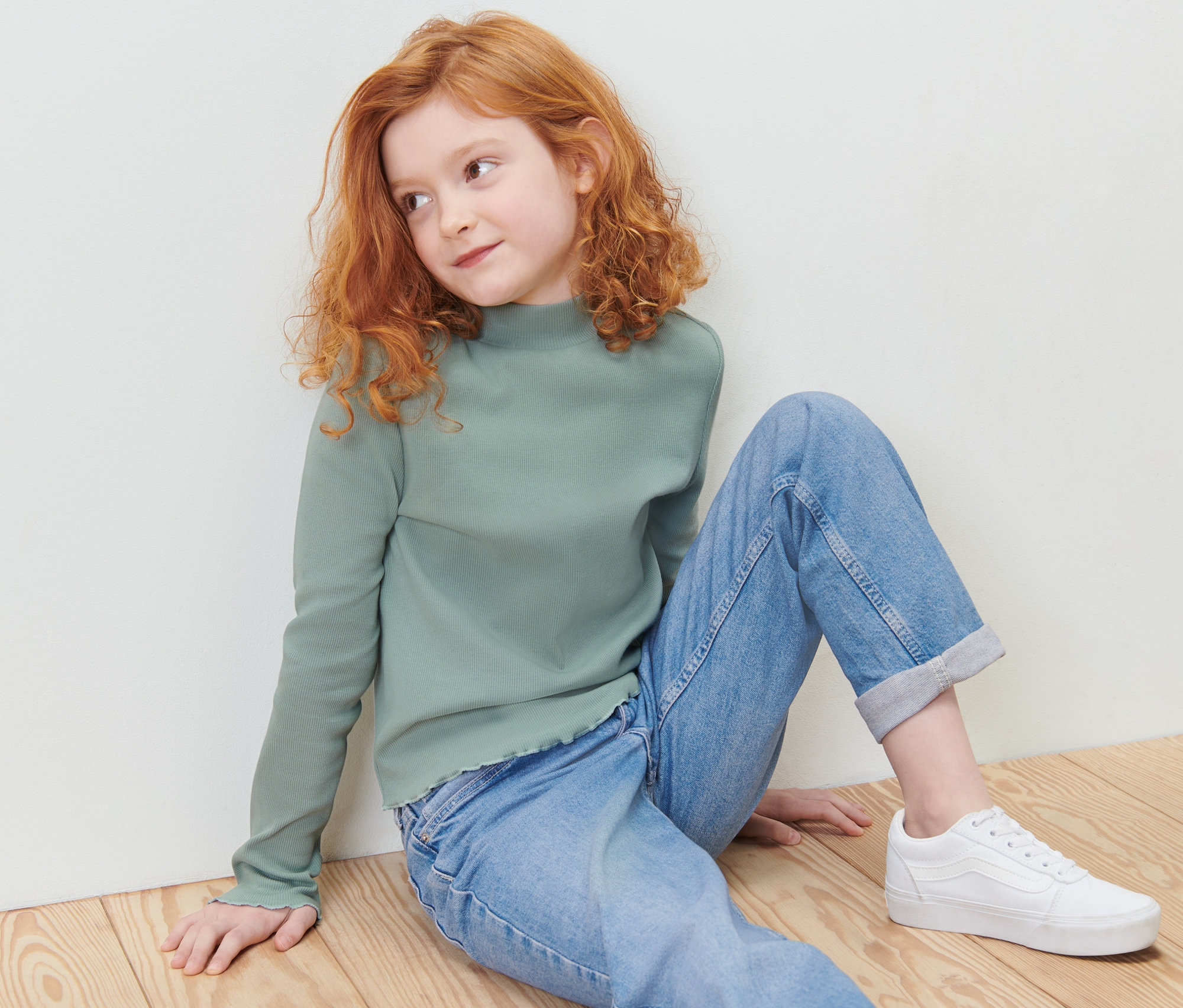 Dievča sedí na podlahe v zelenom tričku s dlhými rukávmi a tregginsách.