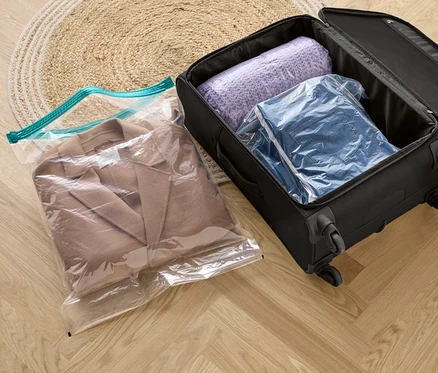 Béžový kabát vo vákuovom cestovnom vrecku, vedľa čierneho kufra s fialovým svetrom a džínsovou bundou vo vákuových cestovných vreckách.