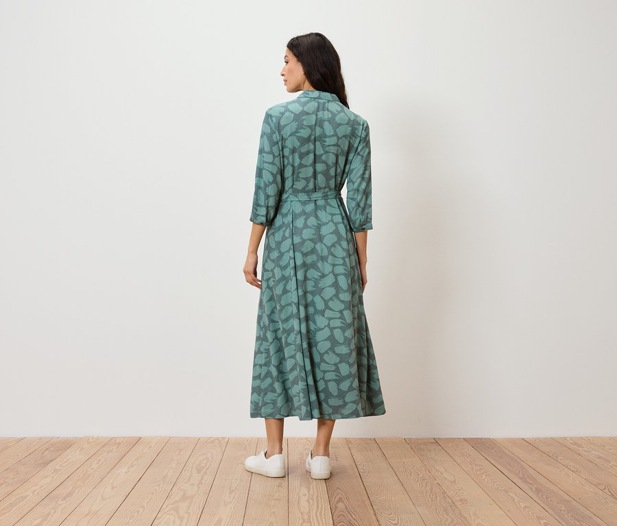 Žena stojí chrbtom k fotoaparátu a má na sebe zelené vzorované šaty a biele tenisky.