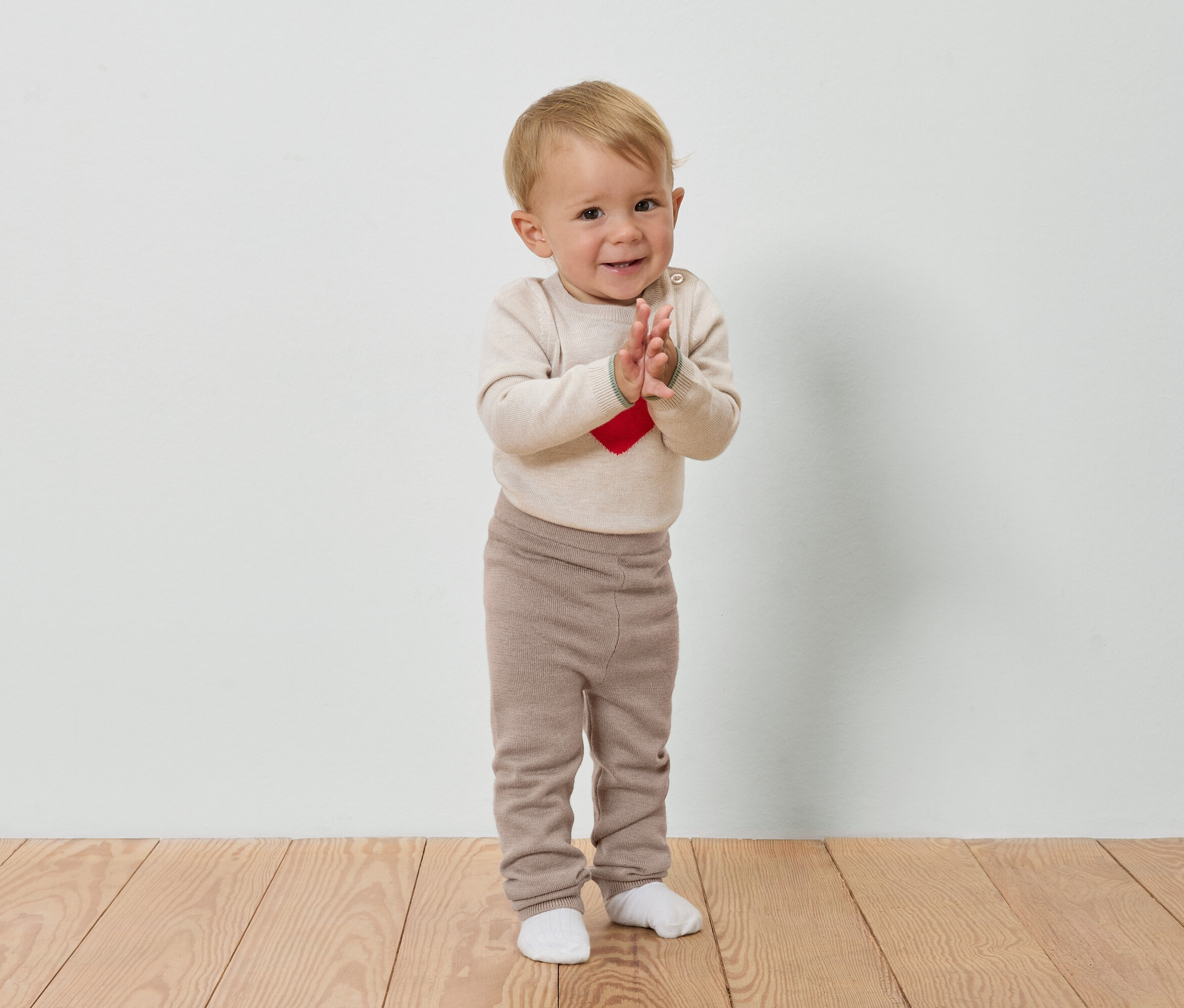 Dieťa s blond vlasmi tlieska rukami na drevenej podlahe pred bielou stenou.