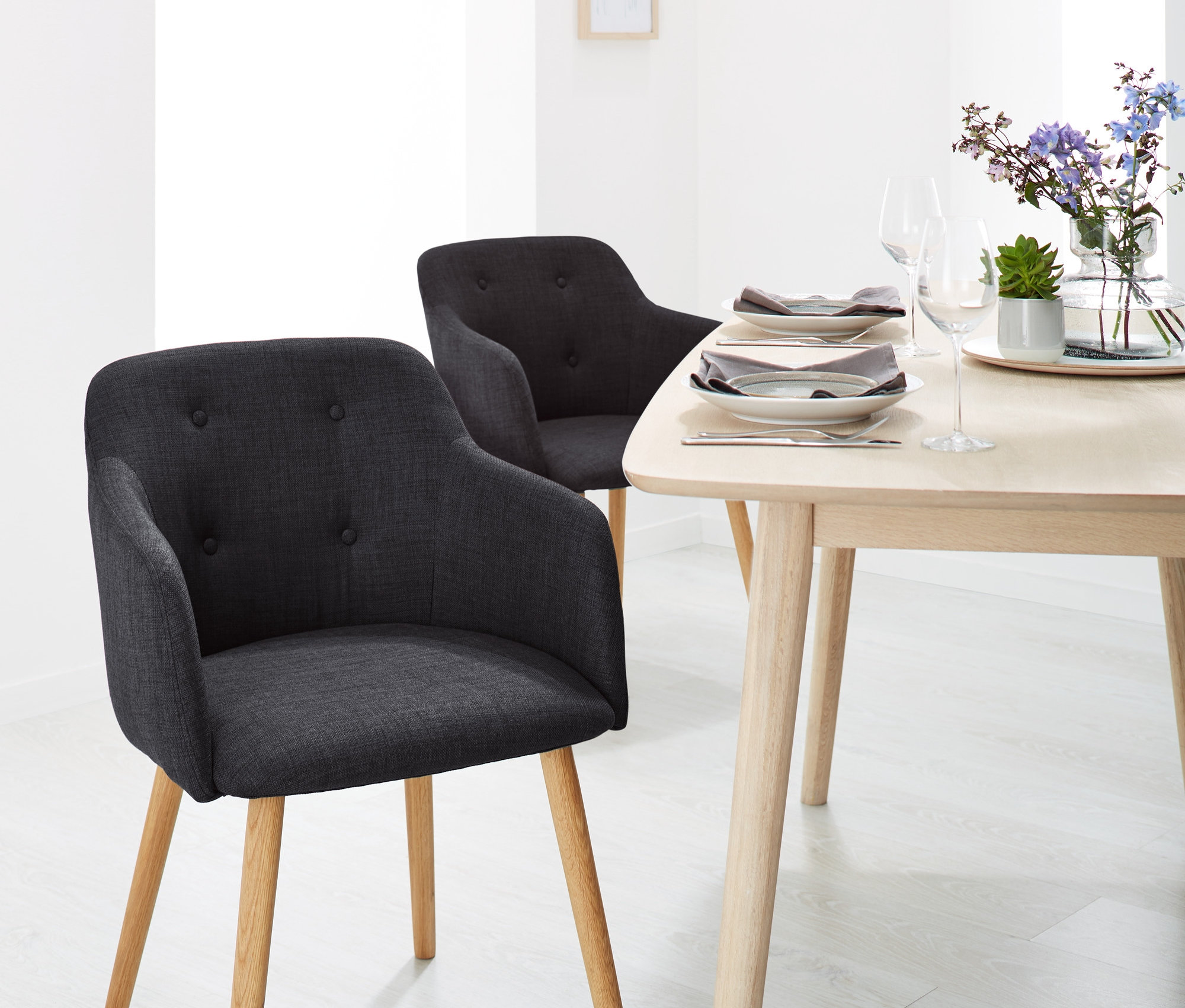 Dve antracitové čalúnené stoličky s opierkami na ruky a drevený stôl s kvetmi a taniermi v jedálni.