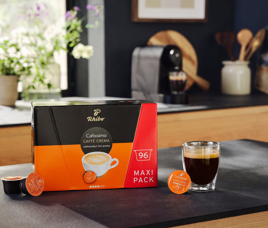 Balenie Cafissimo Caffè Crema vollmundig 96 kapsúl stojí vedľa pohára s kávou. V pozadí je kávovar Cafissimo pure+ black.