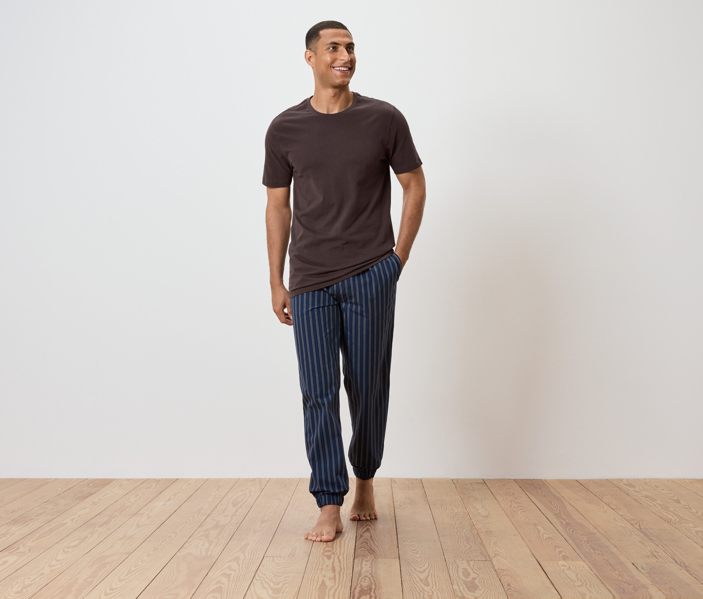 Muž kráča po drevenej podlahe v hnedom tričku a modrých pruhovaných pyžamových nohaviciach.