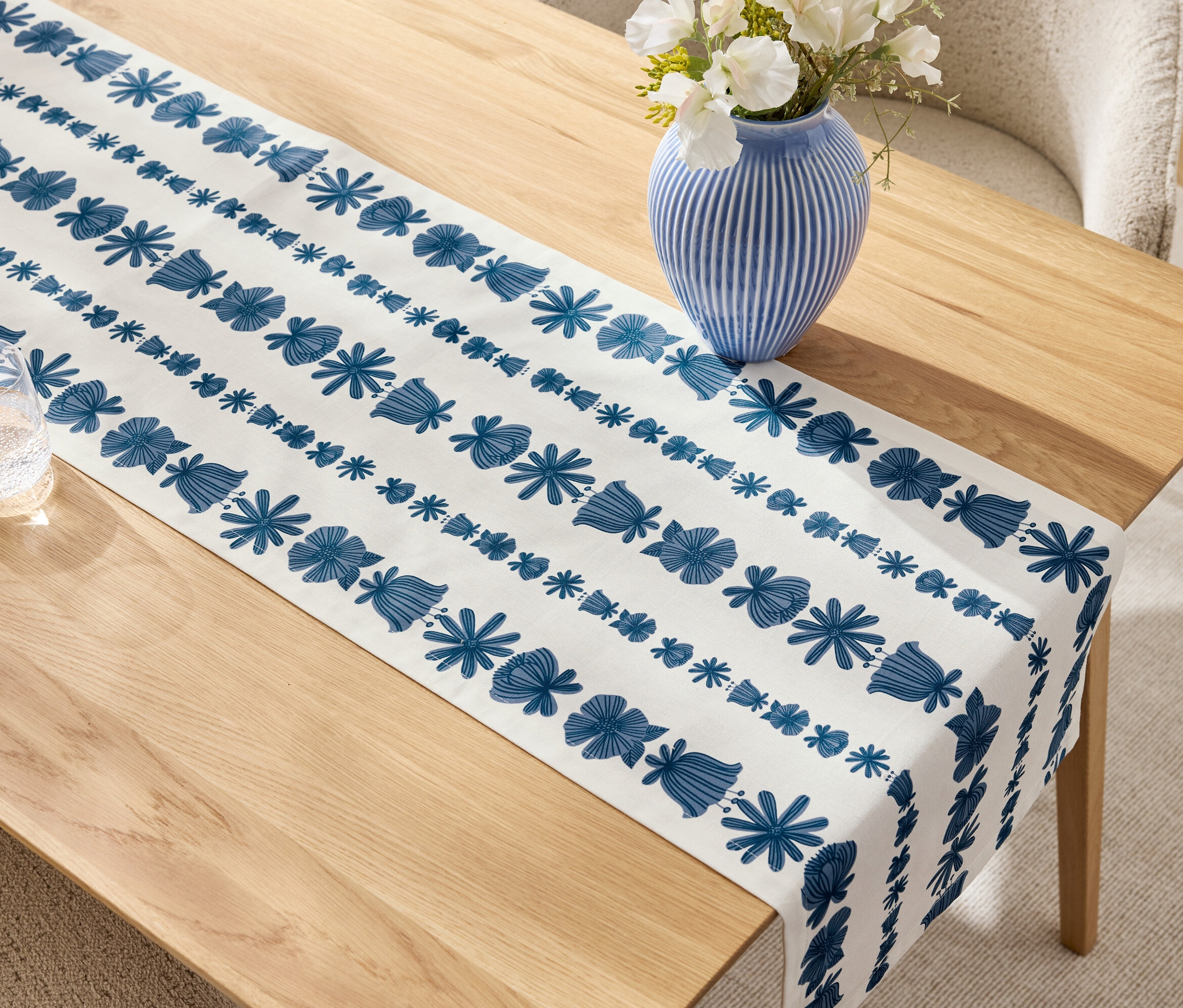 Béžový drevený stôl s modro-bielym behúňom, modrá váza s bielymi kvetmi a pohár.