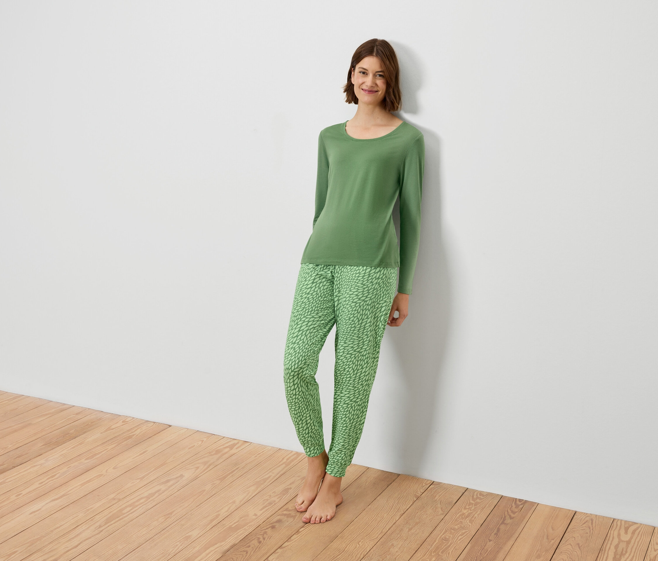 Žena v zelenom pyžame sa opiera o bielu stenu.