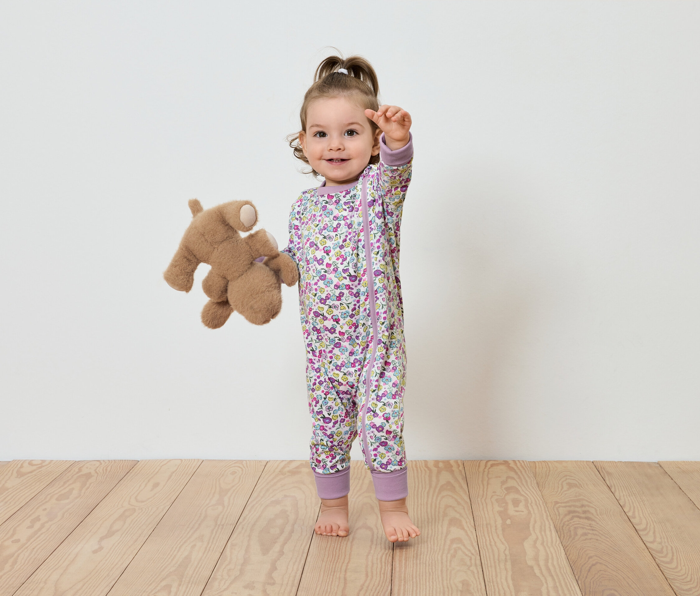 Dieťa v kvetinovom overale stojí s plyšovým medvedíkom.