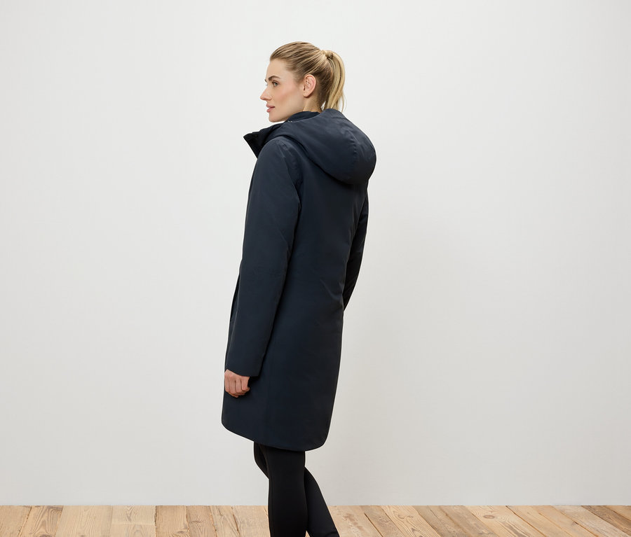 Bočný pohľad na ženu s blond vlasmi stiahnutými do copu, oblečenú v dlhom tmavomodrom kabáte s kapucňou a čiernych legínach.