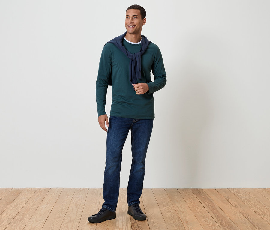 Muž s tmavými vlasmi a úsmevom stojí na drevenej podlahe. Má na sebe zelené tričko s dlhým rukávom, modré džínsy a čierne topánky. Okolo krku má prehodený tmavomodrý sveter.