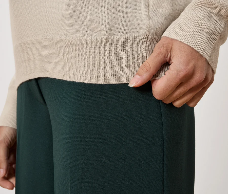 Detailný záber osoby v béžovom svetri a tmavozelených nohaviciach.