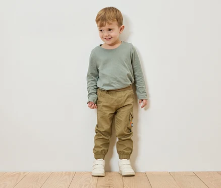 Chlapec s blond vlasmi stojí pred bielou stenou a má na sebe zelené tričko s dlhým rukávom, khaki nohavice a biele topánky.